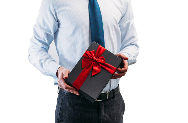 Obchodník drží dárek s červenou stuhou. Chlápek na bílém pozadí v lehké košili s kravatou. Černá dárková krabička v rukou. - Fotografie, Obrázek