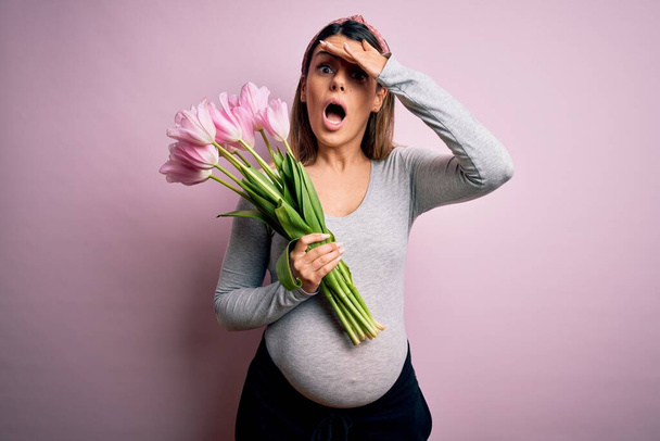 Joven hermosa morena embarazada esperando bebé sosteniendo ramo de tulipanes estresados con la mano en la cabeza, conmocionados con la vergüenza y la cara sorpresa, enojados y frustrados. Miedo y malestar por error
. - Foto, Imagen