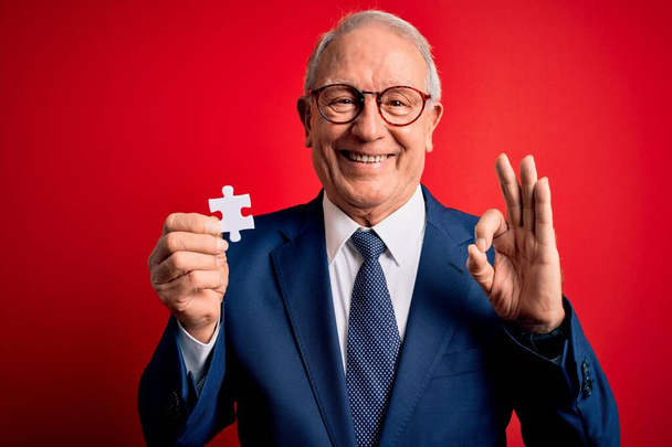 Šedovlasý starší obchodní muž držící puzzle jako odborů a spolupráce dělá v pořádku znamení s prsty, vynikající symbol - Fotografie, Obrázek