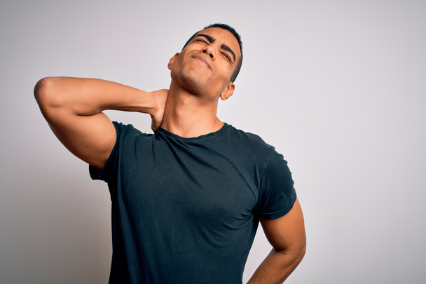Mladý pohledný africký Američan muž na sobě ležérní tričko stojící nad bílým pozadím Utrpení bolesti krku zranění, dotýkání krku s rukou, svalová bolest - Fotografie, Obrázek