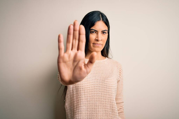 Young beautiful hispanic woman wearing elegant pink sweater over isolated background doing stop sing with palm of the hand. Warning expression with negative and serious gesture on the face. - Foto, Imagem
