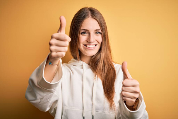 Young beautiful redhead sporty woman wearing sweatshirt over isolated yellow background approving doing positive gesture with hand, thumbs up smiling and happy for success. Winner gesture. - Valokuva, kuva