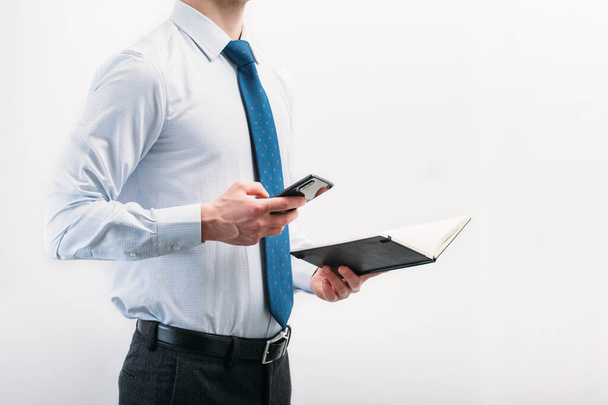 Un gars en chemise légère avec une cravate regarde le téléphone tenant un cahier noir dans sa main sur un fond blanc. Notes électroniques et écrites. - Photo, image