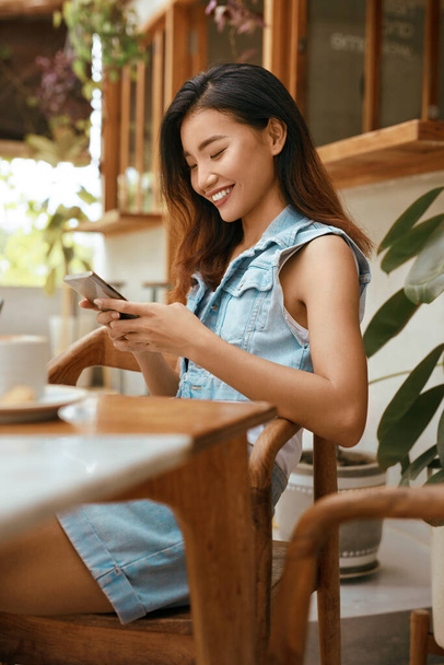 Woman At Cafe With Smartphone. Asian Girl In Casual Clothes Using Mobile Phone For Chatting With Somebody. Digital Technologies For Comfortable Life On Vacation And Successful Business. - Foto, Imagen