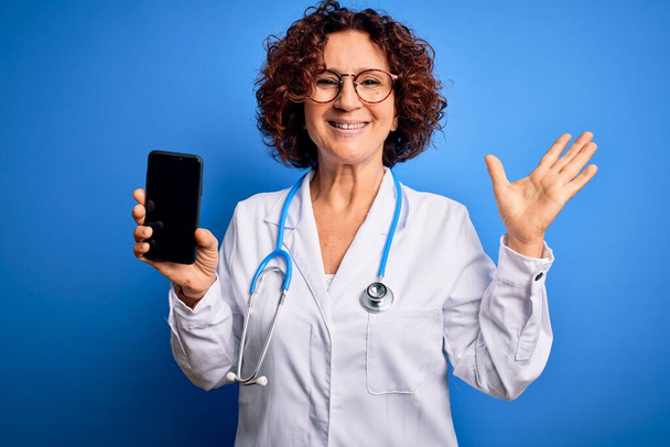 Médico de mediana edad mujer con abrigo y estetoscopio sosteniendo teléfono inteligente mostrando la pantalla muy feliz y emocionada, la expresión ganadora celebrando la victoria gritando con una gran sonrisa y las manos levantadas
 - Foto, imagen
