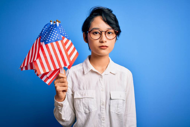 Νεαρό όμορφο ασιατικό πατριωτικό κορίτσι κρατώντας σημαίες των Ηνωμένων Πολιτειών γιορτάζει την ημέρα της ανεξαρτησίας με αυτοπεποίθηση έκφραση στο έξυπνο πρόσωπο σκέφτεται σοβαρά - Φωτογραφία, εικόνα