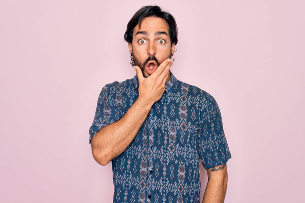 Joven hombre bohemio hispano guapo usando estilo hippie sobre fondo rosa que parece fascinado con la incredulidad, sorpresa y expresión asombrada con las manos en la barbilla
 - Foto, imagen