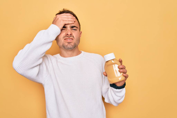 Young handsome man holding bottle of mustard sauce condiment over yellow background stressed with hand on head, shocked with shame and surprise face, angry and frustrated. Fear and upset for mistake. - 写真・画像