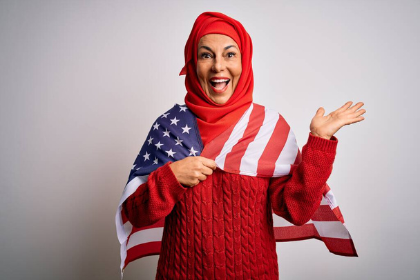 Mujer patriótica de mediana edad vistiendo el hiyab musulmán tradicional sosteniendo la bandera de los estados unidos muy feliz y emocionada, expresión ganadora celebrando la victoria gritando con una gran sonrisa y levantando las manos
 - Foto, imagen
