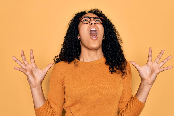 Mujer afroamericana joven con suéter casual y gafas sobre fondo amarillo loco y loco gritando y gritando con expresión agresiva y los brazos levantados. Concepto de frustración
. - Foto, imagen