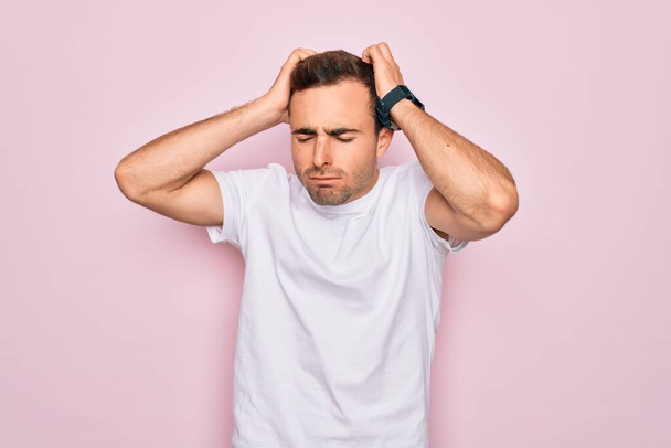 Jóképű, kék szemű férfi alkalmi fehér pólóban, rózsaszín háttér felett állva, fejfájástól szenvedve, kétségbeesetten és stresszesen, mert fájdalom és migrén. Kezeket a fejre!. - Fotó, kép