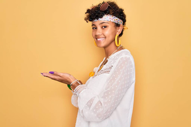Young beautiful african american afro hippie woman wearing sunglasses and accessories pointing aside with hands open palms showing copy space, presenting advertisement smiling excited happy - Photo, image