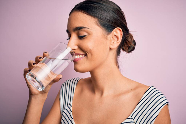 Νεαρή όμορφη μελαχρινή γυναίκα χαμογελά ευτυχισμένη και σίγουρη. Στέκεται με χαμόγελο στο πρόσωπο πίνοντας ένα ποτήρι υγιεινό νερό για να δροσιστεί σε απομονωμένο ροζ φόντο - Φωτογραφία, εικόνα