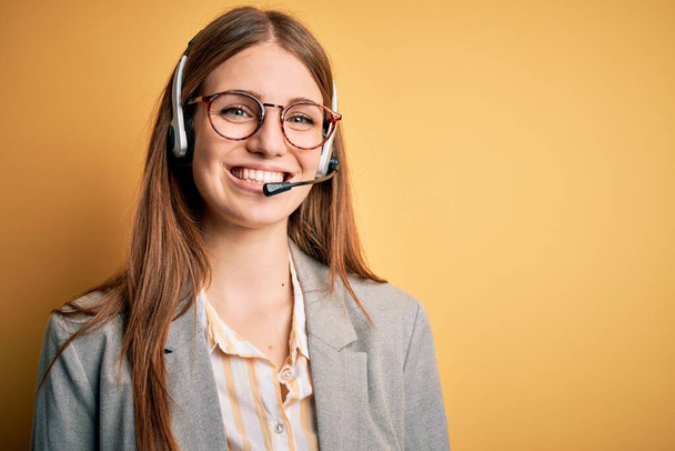 Nuori punapää Call Center agentti nainen ylityöllistetty yllään lasit käyttäen kuulokkeet onnellinen ja viileä hymy kasvoilla. Onnekas henkilö. - Valokuva, kuva