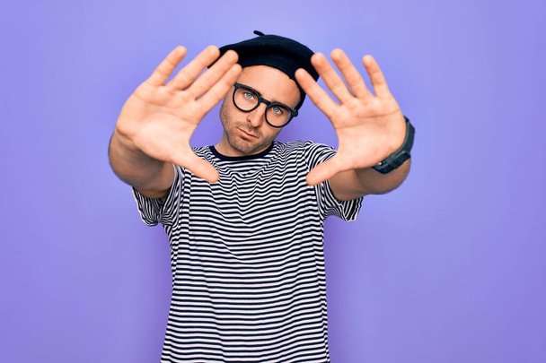 Beau homme aux yeux bleus portant un t-shirt rayé et un béret français sur fond violet faisant le cadre à l'aide des mains, des paumes et des doigts, perspective de la caméra - Photo, image