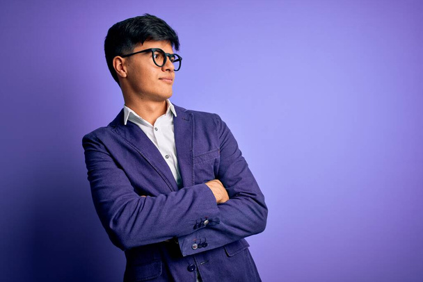 Fiatal jóképű üzletember visel kabát és szemüveg felett elszigetelt lila háttér néz oldalra karok keresztbe meggyőzött és magabiztos - Fotó, kép