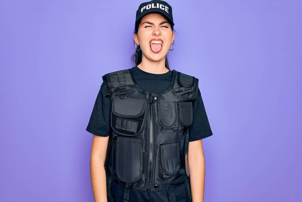 Egy fiatal rendőrnő golyóálló mellényt visel lila háttér fölött, és vicces arckifejezéssel kidugja a nyelvét. Érzelmi koncepció. - Fotó, kép