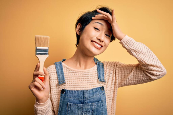 Young beautiful chinese woman painting holding paint brush over isolated yellow background stressed with hand on head, shocked with shame and surprise face, angry and frustrated. Fear and upset for mistake. - Foto, immagini