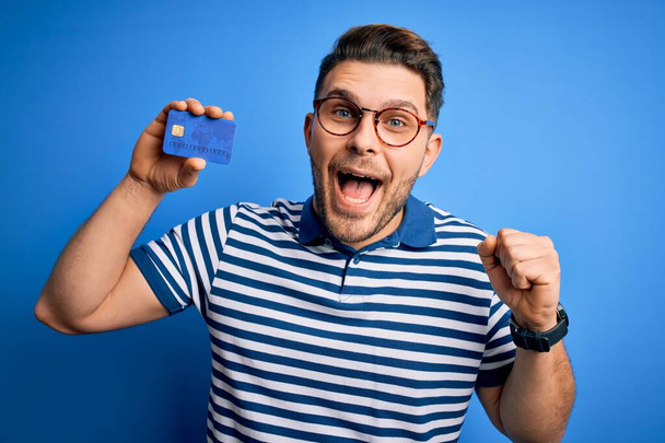 Young man with blue eyes wearing glasses and holding credit card over blue background screaming proud and celebrating victory and success very excited, cheering emotion - 写真・画像