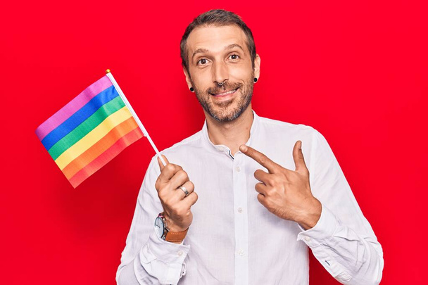 Joven hombre guapo sosteniendo arco iris lgbtq bandera sonriendo feliz señalando con la mano y el dedo
  - Foto, imagen