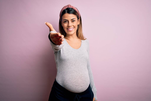 Joven hermosa mujer morena embarazada esperando bebé sobre fondo rosa aislado sonriendo amistoso ofreciendo apretón de manos como saludo y bienvenida. Negocios exitosos
. - Foto, Imagen