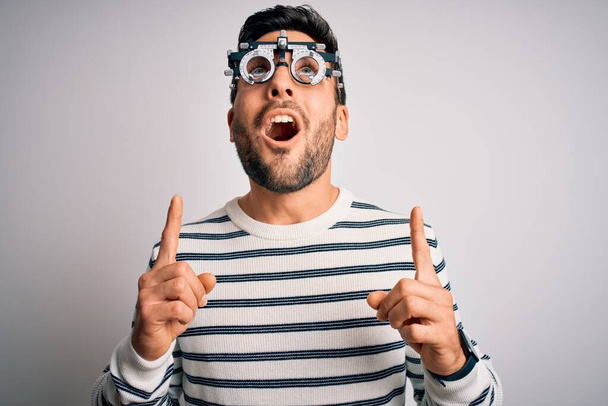 Jonge knappe man met baard draagt optometrie bril over geïsoleerde witte achtergrond verbaasd en verrast omhoog te kijken en te wijzen met vingers en opgeheven armen. - Foto, afbeelding