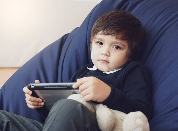Cute maluch chłopiec noszenie munduru szkolnego leżącego na niebieskiej fasoli torba krzesło oglądanie tabletki z misia w pokoju dziecka, Kid i koncepcji technologii - Zdjęcie, obraz