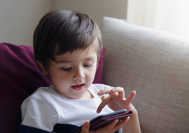 Boldog kisgyermek fiú szórakozás játék mobiltelefon, óvodás gyerek ül a kanapén mosolygós arc néz rajzfilm okos telefon, gyermek használja a mobiltelefon, miközben pihentető otthon. - Fotó, kép