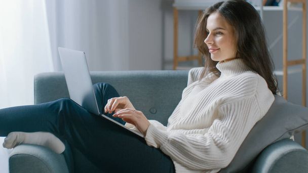 Beautiful Young Woman Works on Laptop Computer while Sitting on the Chair. Sensual Girl Wearing Sweater Works On Notebook Studies, Surfs Internet, Uses Social Media while Relaxing in Cozy Apartment - Fotografie, Obrázek