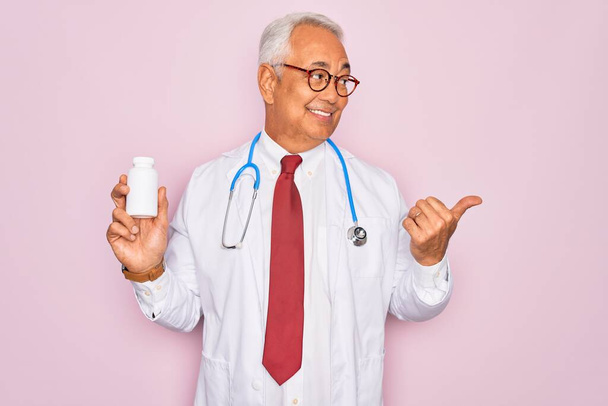 Středního věku senior šedovlasý lékař muž drží léky na předpis přes růžové pozadí ukazuje palcem nahoru na stranu se šťastnou tváří usmívá - Fotografie, Obrázek