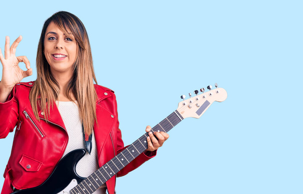 Junge schöne Frau spielt E-Gitarre macht ok Zeichen mit den Fingern, lächelt freundlich gestikulierend hervorragendes Symbol  - Foto, Bild