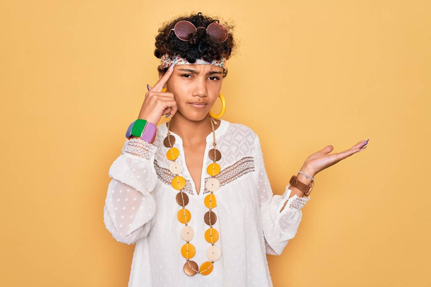 サングラスやアクセサリーを身に着けている若い美しいアフリカ系アメリカ人のアフロヒッピー女性は、コピースペースを示し、額に指を指すことで混乱し、いらいらしています。考えてみて. - 写真・画像