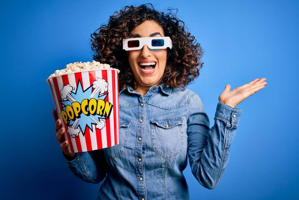 Joven hermosa mujer árabe rizado viendo la película usando gafas 3d comer bocadillo de palomitas de maíz muy feliz y emocionado, expresión ganadora celebrando la victoria gritando con una gran sonrisa y las manos levantadas
 - Foto, imagen