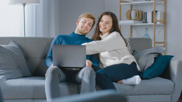 Cute Young Couple Use Laptop Computer, Have Fun, Laughs, while Sitting on the Couch in the Cozy Apartment. Couple Shopping on Internet, Using Social Media, Watching Videos and Streaming Content - Φωτογραφία, εικόνα