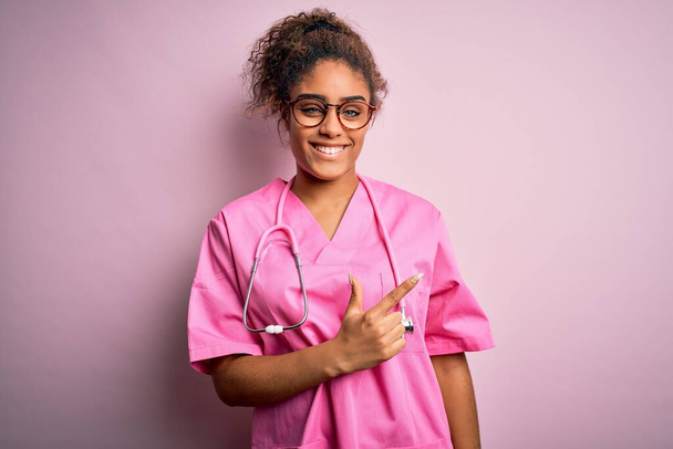 Африканська медсестра з Америки, одягнена в медичну форму і стетоскоп над рожевим фоном весела з посмішкою, що вказує пальцем на бік з радісним і природним виразом обличчя - Фото, зображення
