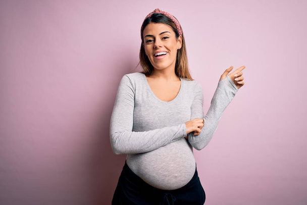 Junge schöne brünette Frau schwanger Baby auf isoliertem rosa Hintergrund mit einem breiten Lächeln im Gesicht, zeigt mit der Hand und dem Finger zur Seite in die Kamera. - Foto, Bild
