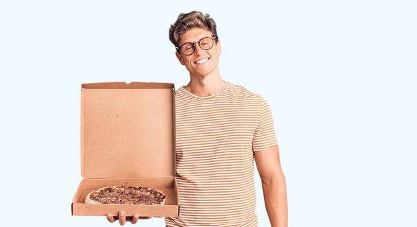 Młody przystojny mężczyzna trzyma pudełko po pizzy patrząc pozytywnie i szczęśliwy stojąc i uśmiechając się z pewnym uśmiechem pokazując zęby  - Zdjęcie, obraz