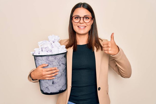 Молодая красивая брюнетка бизнес женщина держит полный ящик бумаги калека бумаги улыбаясь счастливым и позитивным, большой палец вверх делает отлично и знак одобрения
 - Фото, изображение