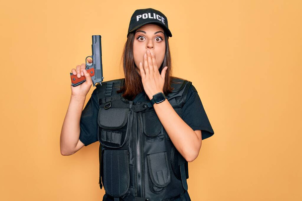 Fiatal gyönyörű barna rendőr nő visel rendőrségi egyenruha golyóálló és sapka segítségével fegyvert fedél száj kézzel sokkolt szégyen a hiba, kifejezés a félelem, félek a csend, titkos koncepció - Fotó, kép