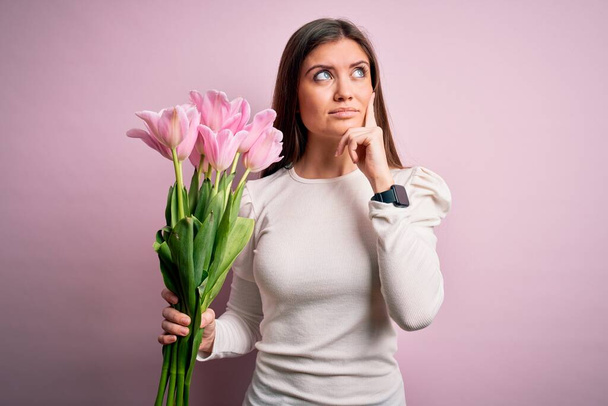 Young beautiful romantic woman with blue eyes holding beauty bouquet of pink tulips serious face thinking about question, very confused idea - Photo, Image