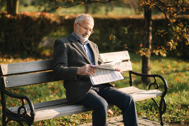 Przystojny dziadek z piękną brodą w szarej kurtce siedzi na ławce w parku i czyta gazetę. Senior siwowłosy mężczyzna w okularach - Zdjęcie, obraz