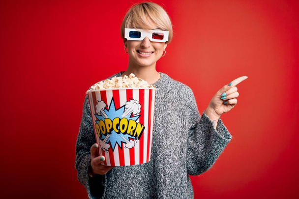 Fiatal szőke nő rövid haj visel 3D-s film szemüveg és eszik popcorn a moziban nagyon boldog mutató kézzel és ujjal oldalra - Fotó, kép