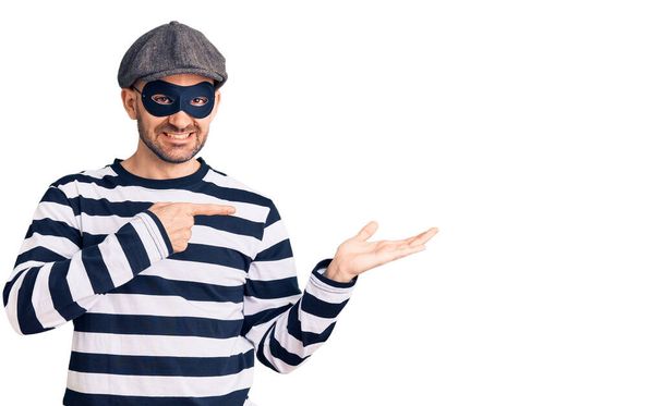 Junger gutaussehender Mann mit Einbrechermaske staunt und lächelt in die Kamera, während er mit der Hand präsentiert und mit dem Finger zeigt.  - Foto, Bild