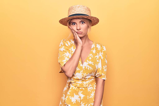 Bella donna bionda in vacanza indossando cappello estivo e vestito su sfondo giallo pensando cercando stanco e annoiato con problemi di depressione con le braccia incrociate. - Foto, immagini