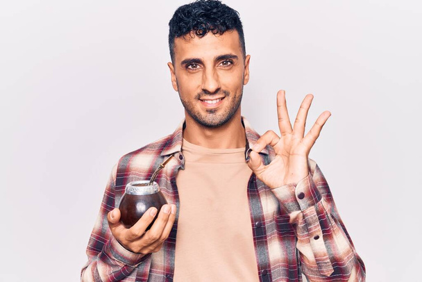 Young hispanic man drinking mate infusion doing ok sign with fingers, smiling friendly gesturing excellent symbol  - Foto, immagini