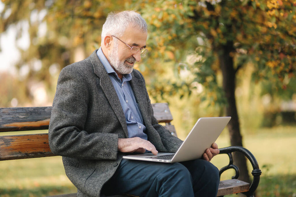 Starszy mężczyzna używa laptopa w parku. Przystojny starszy pan w okularach pracuje na zewnątrz - Zdjęcie, obraz