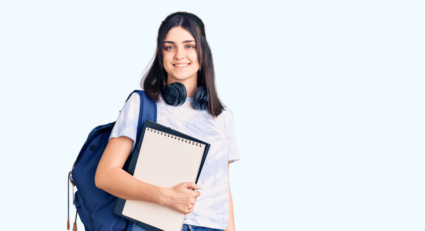 Menina bonita nova usando mochila estudante segurando notebook olhando positivo e feliz de pé e sorrindo com um sorriso confiante mostrando dentes
  - Foto, Imagem