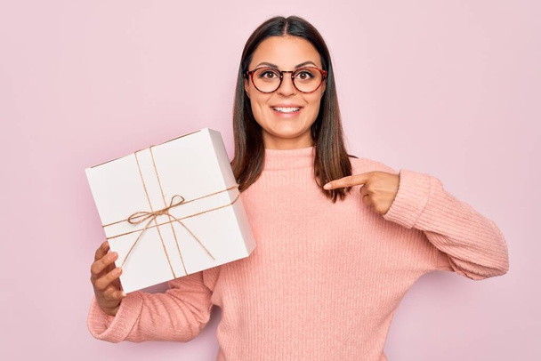 Young beautiful brunette woman holding birthday gift over isolated pink background pointing finger to one self smiling happy and proud - Zdjęcie, obraz