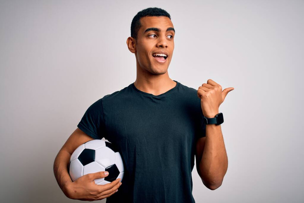 Hombre afroamericano guapo jugando fútbol sosteniendo pelota sobre fondo blanco sonriendo con la cara feliz mirando y señalando hacia el lado con el pulgar hacia arriba
. - Foto, imagen