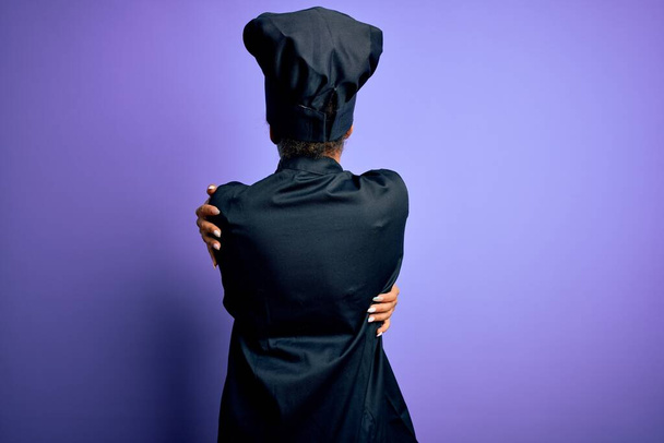Joven cocinera afroamericana con uniforme de cocina y sombrero sobre fondo púrpura Abrazarse feliz y positivo desde atrás. Amor propio y cuidado personal
 - Foto, Imagen